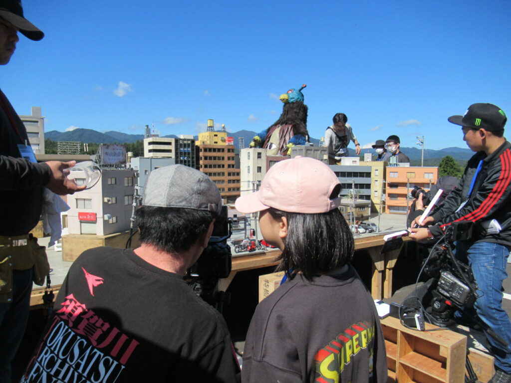 青空の下、精巧に作られたミニチュアの街並みを背景に、大きな一つ目が付いた茶色の怪獣の着ぐるみを囲んでスタッフたちが撮影の準備や調整を行っている、特撮映画のメイキング風景を思わせる臨場感あふれる写真