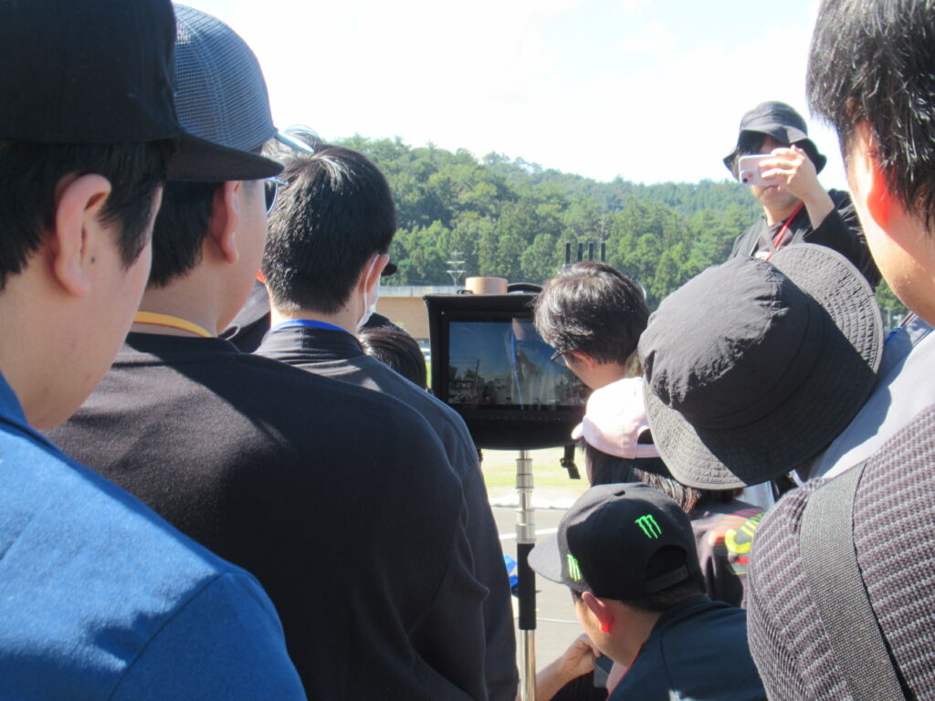 屋外で参加者たちが小型モニターに映る撮影映像を囲み込み真剣に確認している様子を捉えた写真