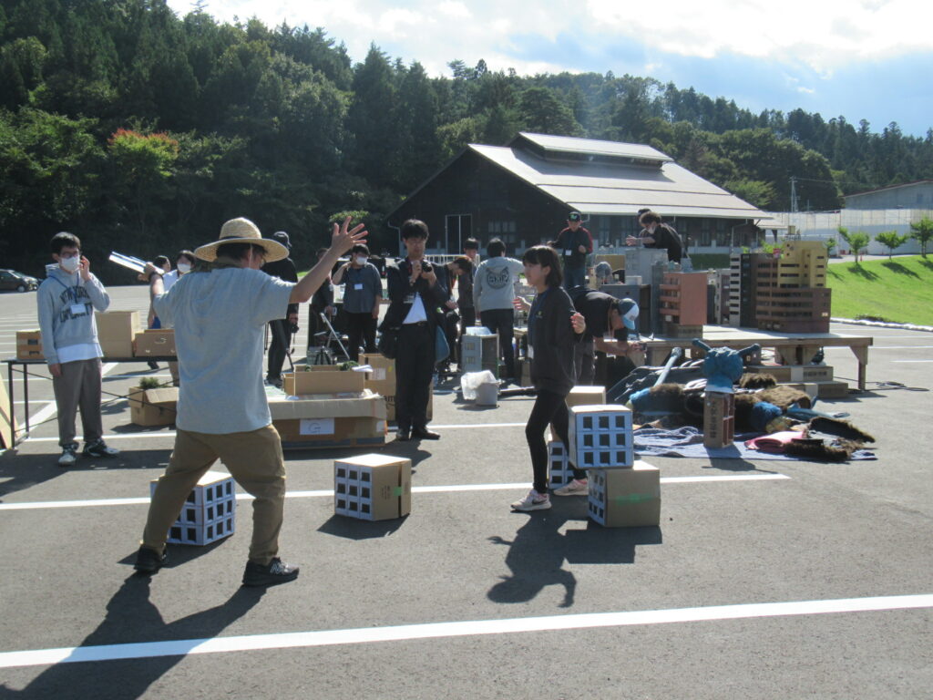 屋外の駐車場で参加者が段ボール製の建物模型や道具を配置しつつ演出指示に応じて動きを確認する様子を捉えた写真