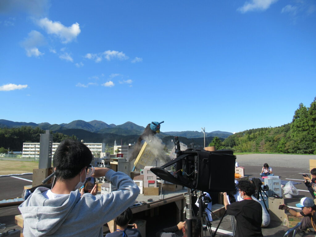 抜けるような青空の下、精巧なミニチュアの街並みの中で怪獣がビルをなぎ倒し土煙が舞う迫力の瞬間を、カメラやスマートフォンを構えた撮影スタッフたちが真剣な眼差しで捉える、特撮映画のメイキング風景を記録した写真