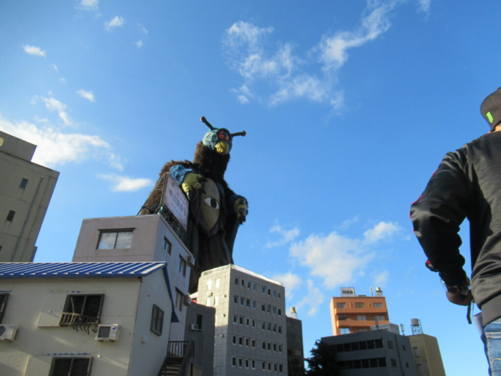 抜けるような青空の下、精巧に作られたミニチュアの街並みの向こうから、青い頭部と一つ目の胴体を持つ巨大な怪獣が姿を現し、ビル群を見下ろすように立っている、特撮映画の劇中シーンを再現したような迫力のある写真