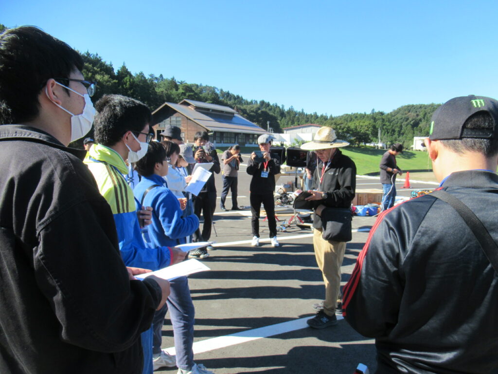 屋外の広場で参加者が書類を手に講師の説明を囲んで真剣に聞き入る様子を捉えた写真