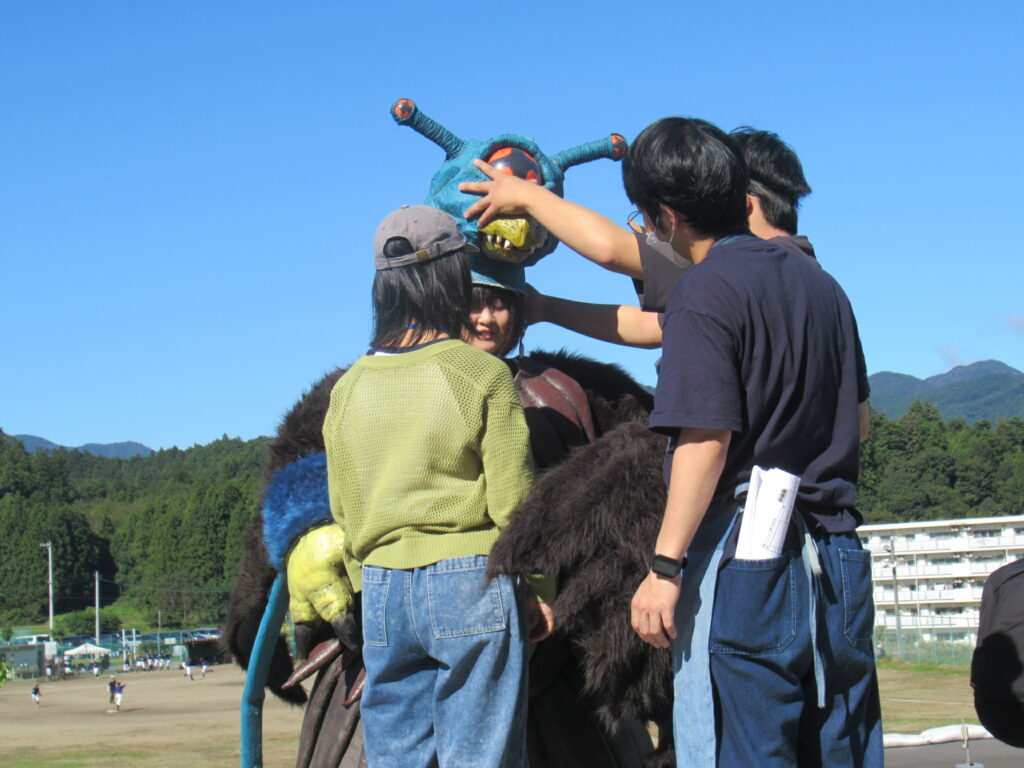 晴天の屋外にて、青い頭部に触角と大きな目が特徴的な怪獣の着ぐるみを着用する女性を、周囲のスタッフたちが笑顔で手伝いながら頭部のパーツを慎重にセットしている、和やかな雰囲気の特撮映画制作現場を捉えた写真