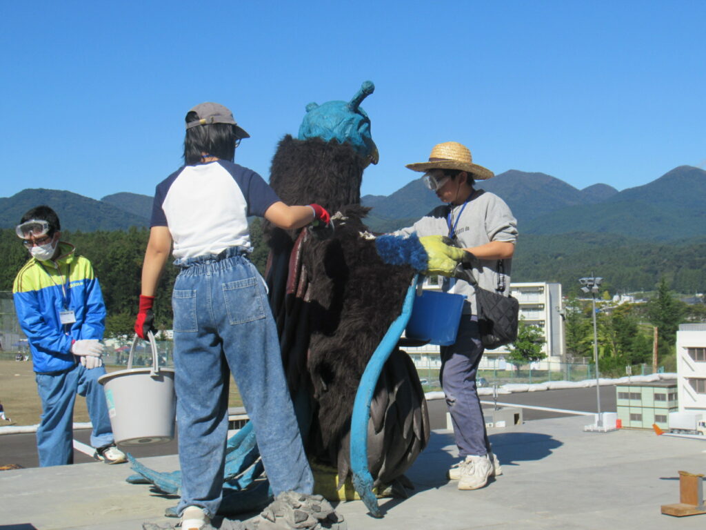 晴天の屋外にて、大きな一つ目が特徴的な怪獣の着ぐるみを囲み、バケツを手にしたスタッフたちが細かな調整やメンテナンスを施している、特撮映画の撮影に向けた準備作業の様子を生き生きと映し出した写真