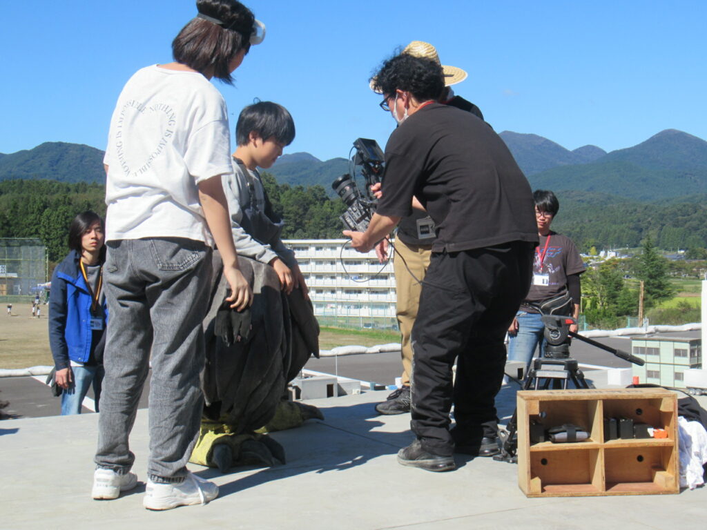 晴天の屋外にて、着ぐるみの足部分のみを装着した少年の姿を、ビデオカメラを構えたスタッフが至近距離から撮影している、特撮映画の制作における演技指導や細部カットの撮影風景を捉えた写真