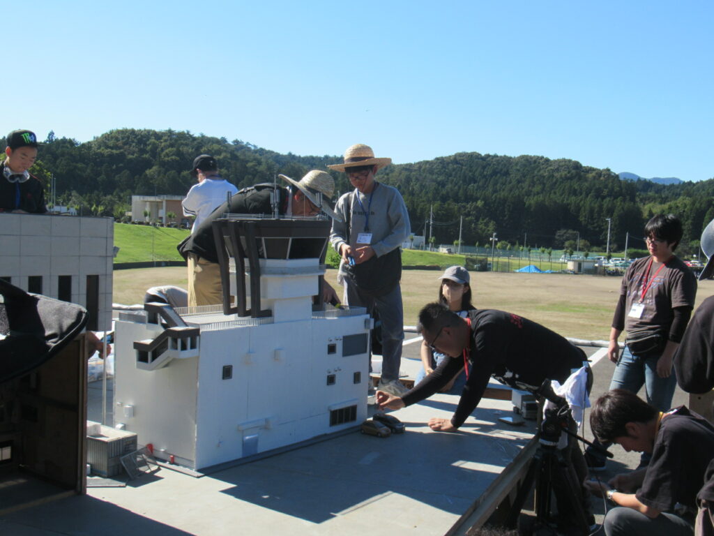 広い青空の下、屋外の広場で人々が複雑な形状の白色大型建物模型と周辺装置を囲み協力して精密な作業を進め、背景には緑の森が広がる様子を写した写真