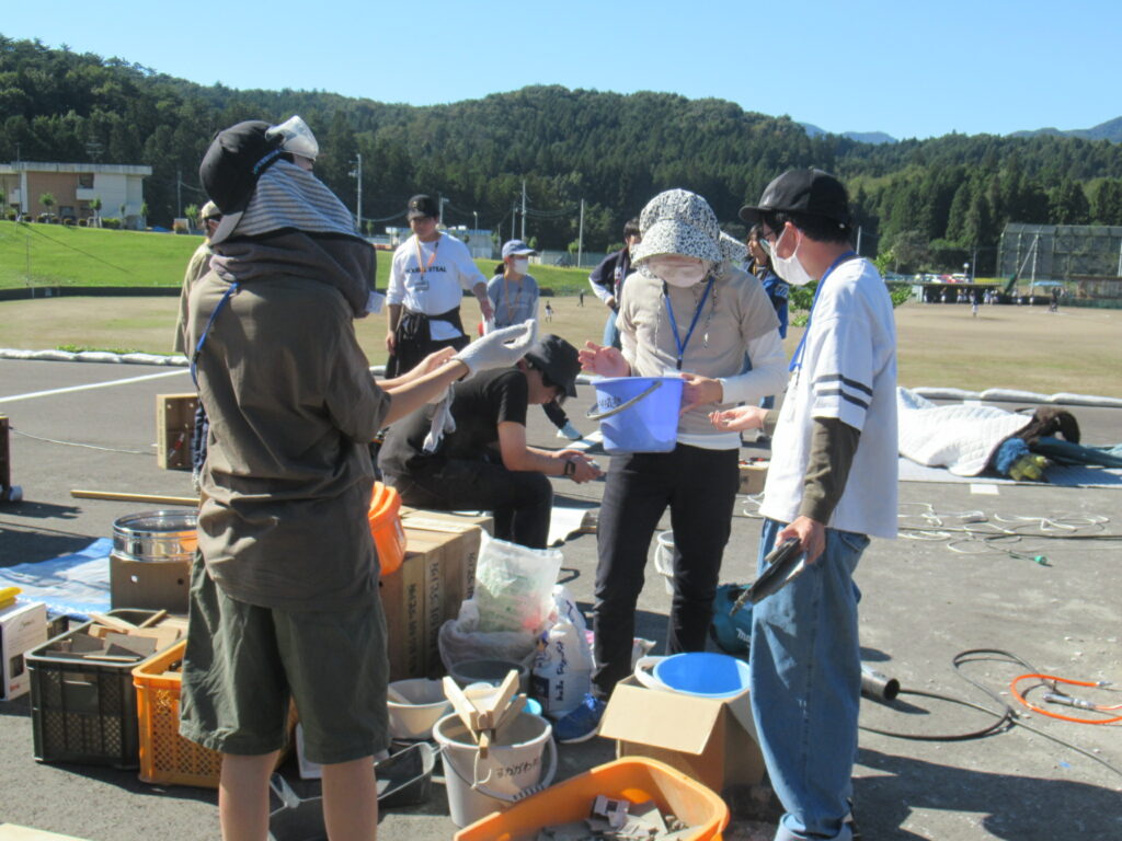 晴天の屋外広場で複数の人々が箱やバケツなど多くの資材を広げ作業内容を相談しながら準備を進めている様子を映した写真