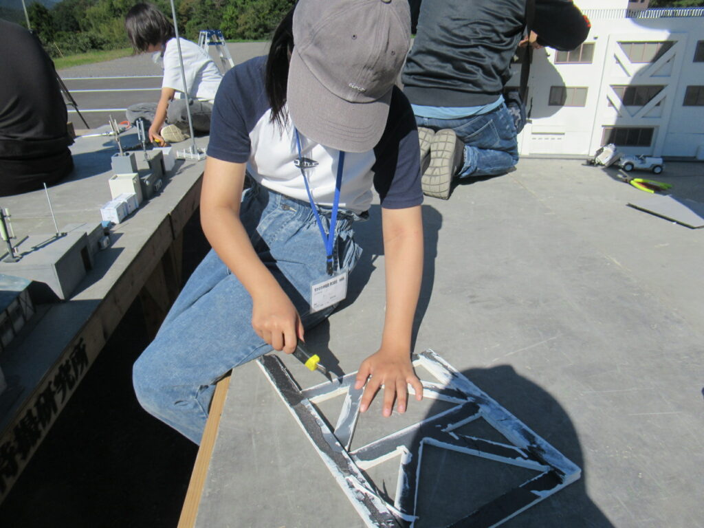 屋外で人物が大型模型の一部を工具で丁寧に補修している様子を日差しの下で捉えた写真