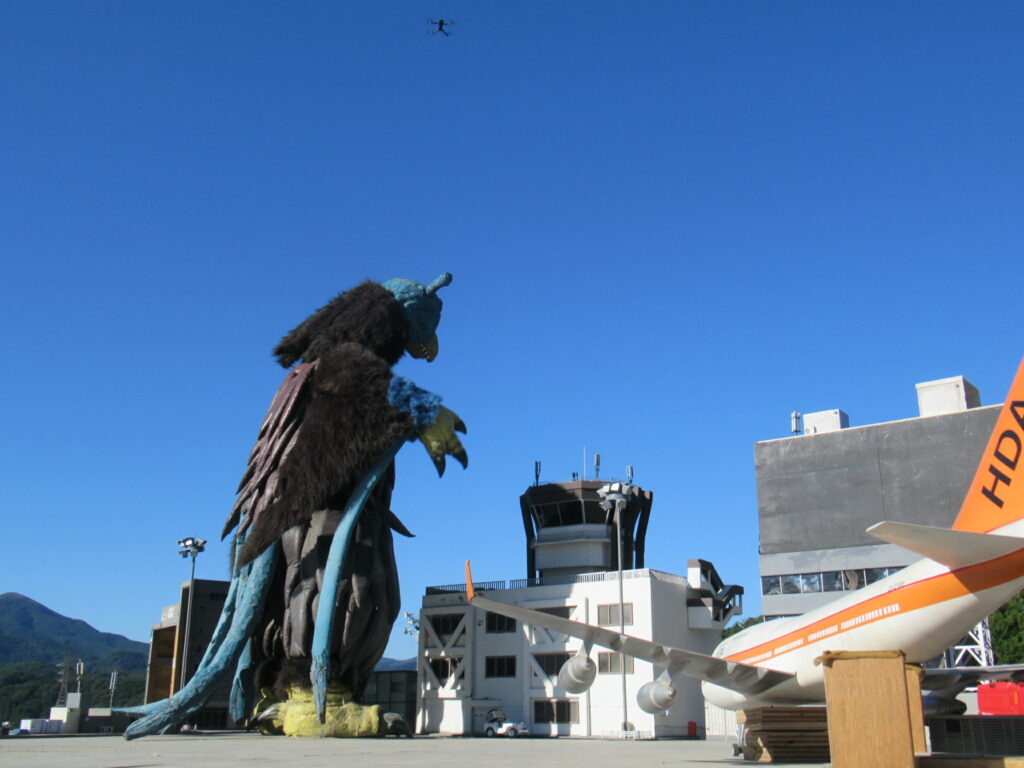 青空の下で巨大な怪獣の着ぐるみが管制塔や航空機模型の前に立ち上空を飛ぶ小型ドローンと共存する様子を捉えた写真