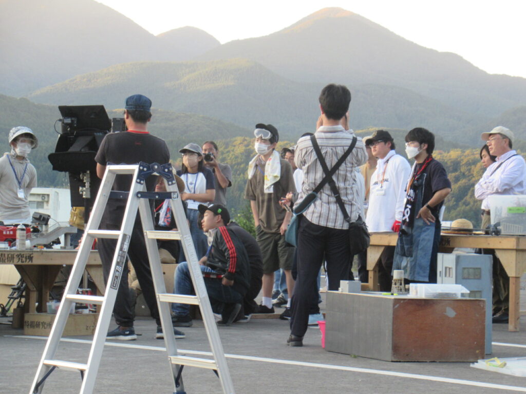 夕暮れの山並みを背景に、屋外で多数の人々が機材や脚立の周囲に集まり作業内容を確認している様子を写した写真