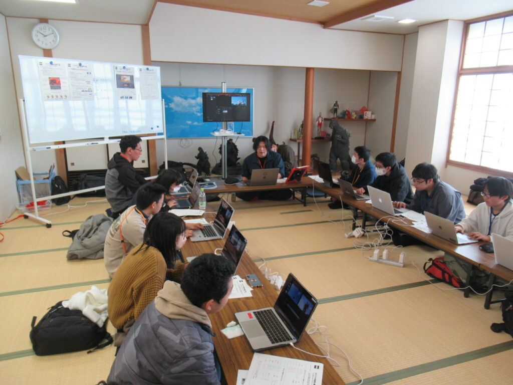 畳の部屋で参加者たちがノートパソコンを広げ白板や大型モニターを囲んで作業に取り組む様子の写真