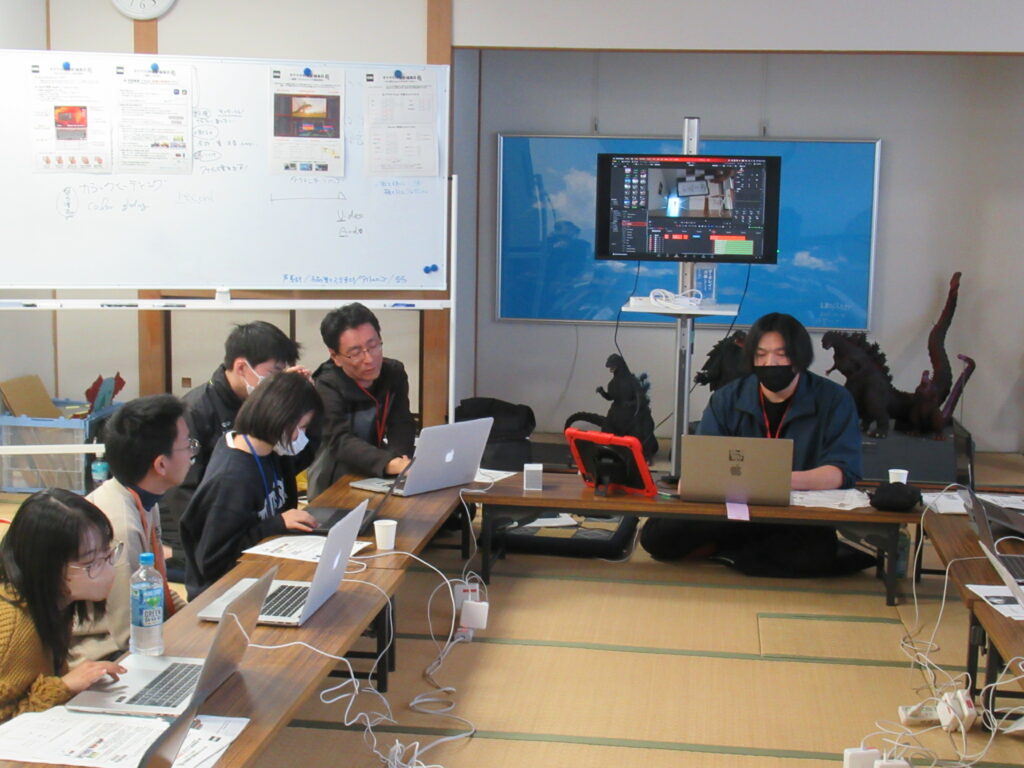 畳敷きの和室で、長机を囲んだ学生たちがノートパソコンを操作しており、講師が画面を覗き込みながらアドバイスを送ったり、大型モニターに編集画面を表示したりして動画制作を学ぶ、特撮映像の仕上げ工程を捉えた写真