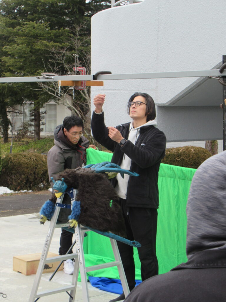 屋外でグリーンスクリーン前に梯子と怪獣風の小道具を配置しスタッフが撮影準備を進める写真