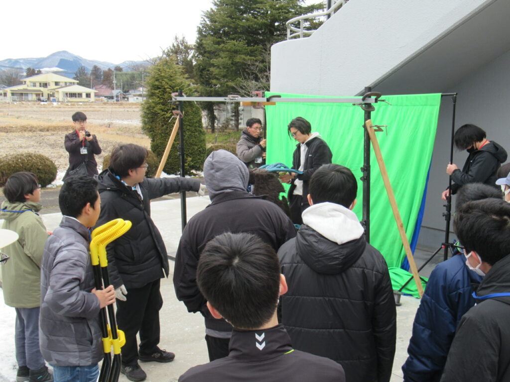 屋外の建物脇で緑色のスクリーンが設置された撮影現場を参加者たちが囲んで準備や指示を交わす写真