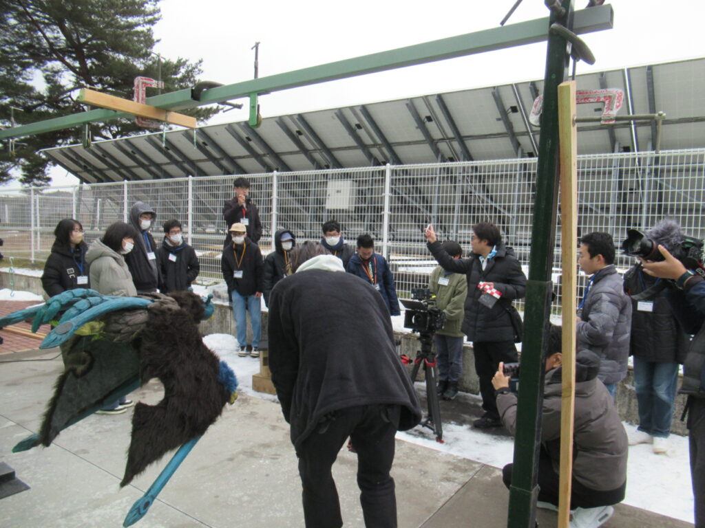 屋外の撮影現場で怪獣風小道具と機材を前に指示を出すスタッフを周囲の参加者が見守る写真