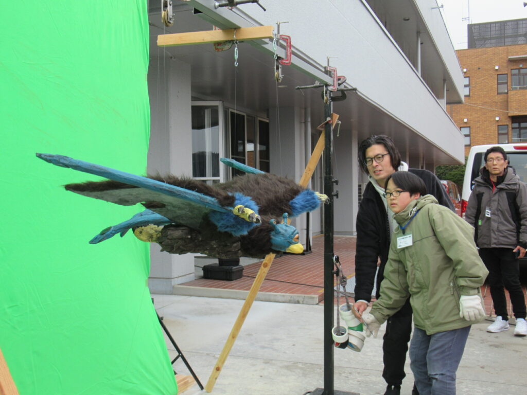 屋外の撮影現場で、緑色のクロマキー用スクリーンの前に吊るされた毛むくじゃらの飛行怪獣のプロップ（小道具）を、講師と学生が真剣な表情で操りながら動きの確認を行っている、特撮映像の撮影風景を捉えた写真