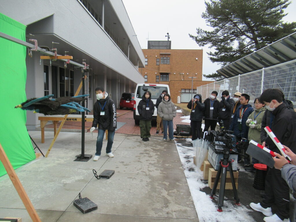 屋外の撮影現場で、緑色のクロマキー用スクリーンの前に吊るされた毛むくじゃらの飛行怪獣を、カメラやカチンコを構えた学生たちが真剣な表情で見守りながら撮影を進めている、冬の特撮ワークショップの風景を捉えた写真