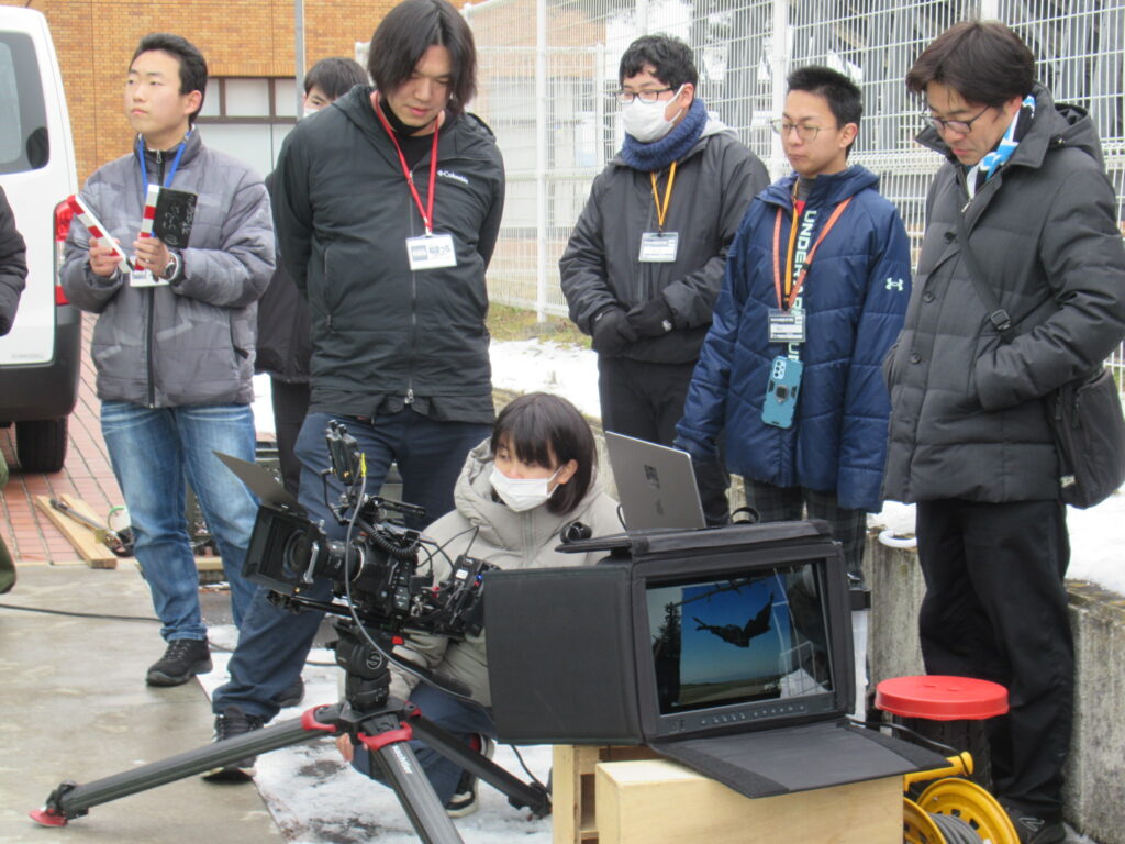 屋外の撮影現場で大型カメラとモニターを前に参加者が映像を確認しつつ機材操作の様子を見守る写真
