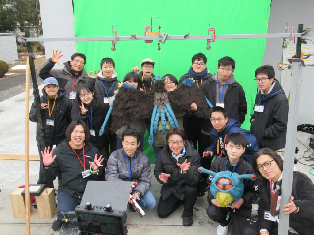 緑色のクロマキー用スクリーンの前に吊るされた飛行怪獣のプロップ（小道具）を囲み、防寒着を着た学生たちや講師が笑顔で怪獣のポーズをとりながら集合写真に収まっている、屋外での特撮映像撮影ワークショップの様子を捉えた写