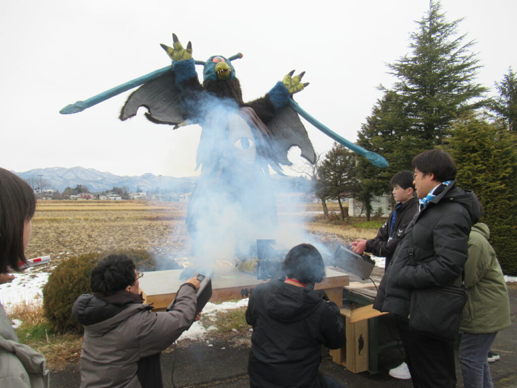 冬の田園地帯に設置された台座の上で、大きな翼を広げた一つ目の怪獣スーツが咆哮するようなポーズをとり、その足元で学生たちがスモークマシンを操作して白い煙を立ち昇らせている、特撮映画のワンシーンのような撮影風景の写真