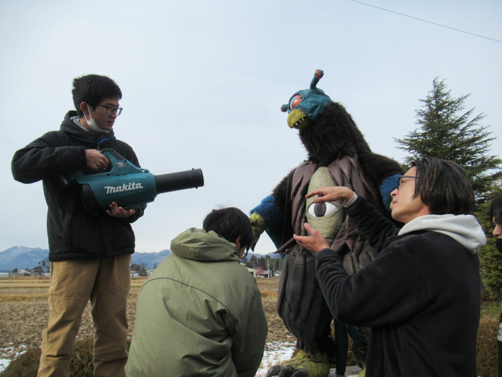 冬の田園地帯で、大きな一つ目の怪獣スーツの前にしゃがみ込むスタッフに対し、左側の男子学生が電動ブロワーを構えて風を送る準備をしており、右側の男性が手で動きの指示を出している、特撮映画の演出風景を捉えた写真