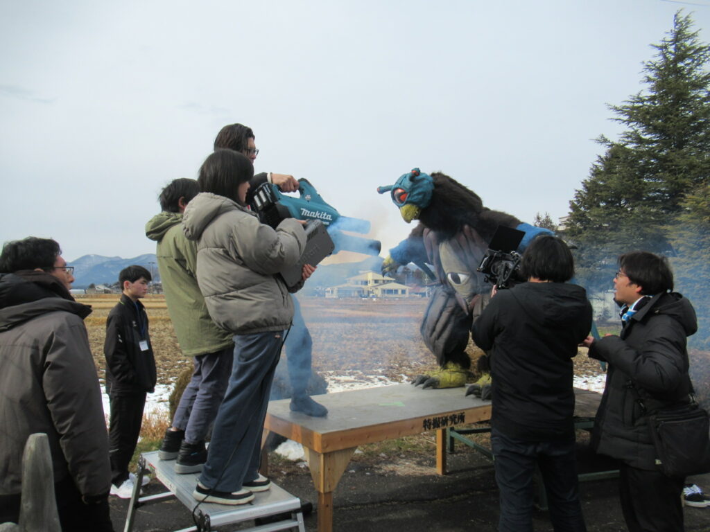冬の田園地帯を背景にした屋外セットで、大きな一つ目の怪獣スーツに向かって、学生たちが電動ブロワーで風を送ったりスモークマシンで煙を発生させたりしながら、カメラを回して特撮シーンを撮影している風景の写真