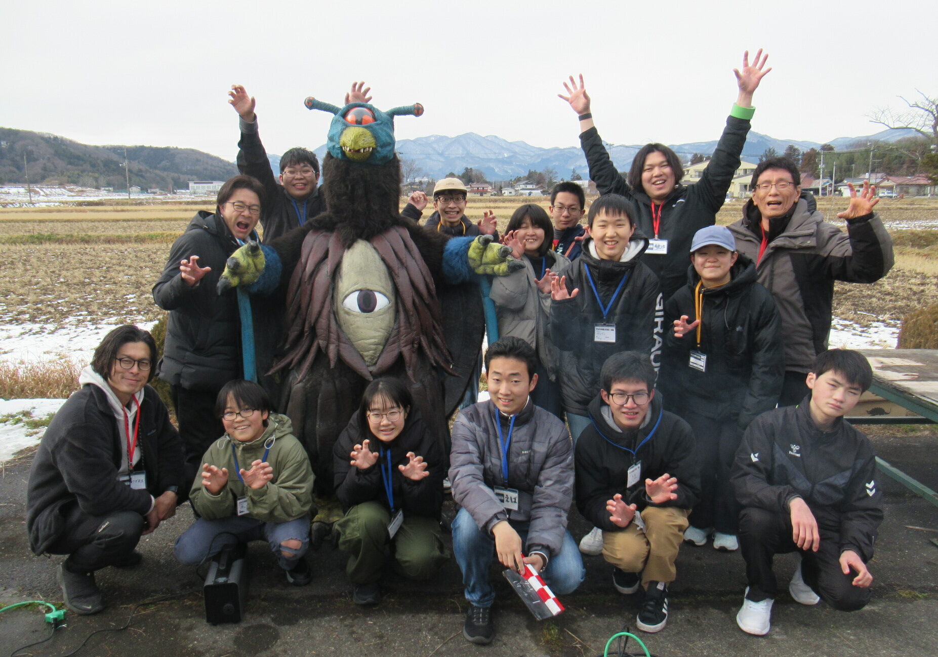 冬の田園風景を背景に、中央に立つ大きな一つ目の怪獣スーツを囲んで、防寒着を着た学生たちや講師が笑顔で怪獣のポーズをとったり手を挙げたりして並んでいる、屋外での特撮ワークショップの記念写真