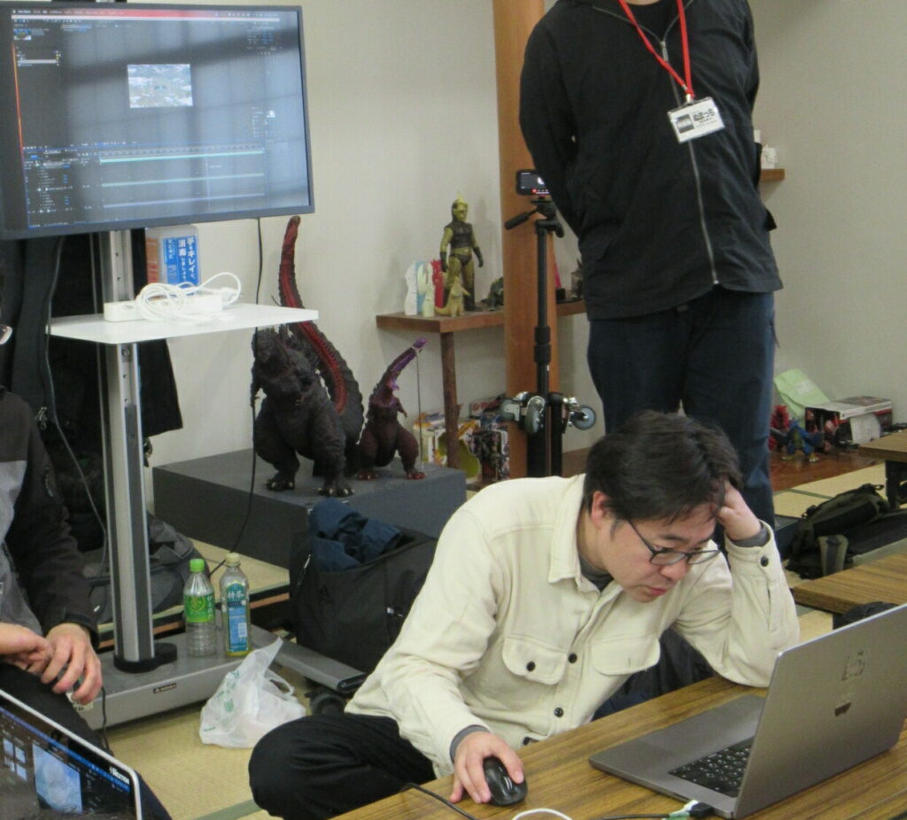 畳敷きの和室で、眼鏡をかけた男性がノートパソコンの画面を険しい表情で見つめながら、マウスを操作して映像編集の仕上げ作業に没頭しており、背後には大型モニターや複数の怪獣フィギュアが置かれている、制作現場の緊迫した一瞬を捉えた写真