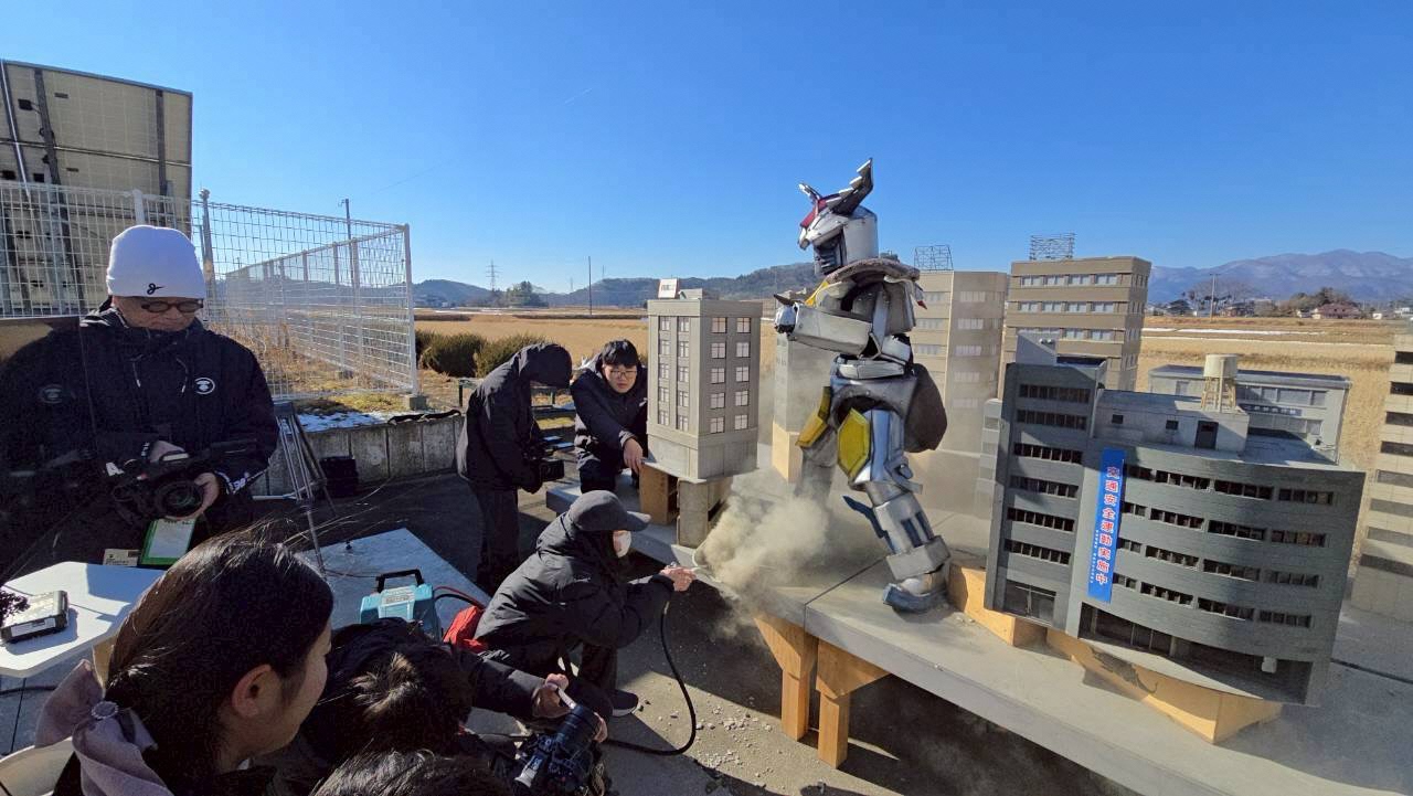 屋外でクワガッシャーの特撮カットを撮影している様子、塾生が土ぼこりを上げる特殊効果を担当したり、カメラを回したり、ロボのスーツを着てアクターを務めたりしている