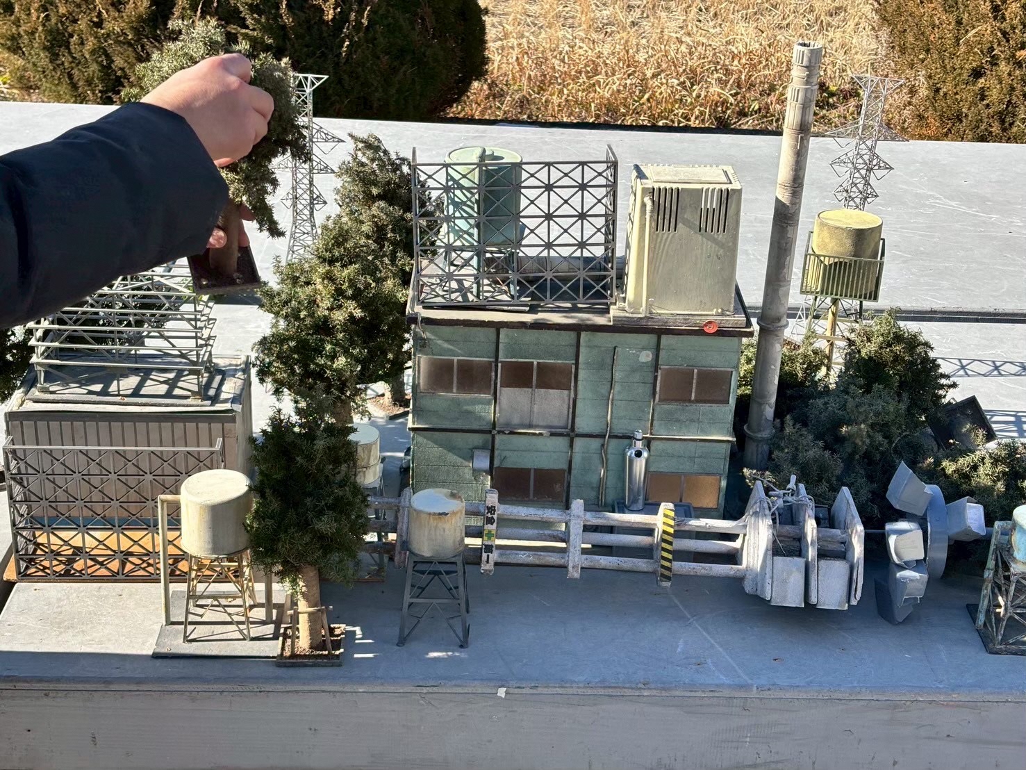 屋外で特撮カットの撮影準備をしてる様子、建物のミニチュアが平台に置かれており、木のミニチュアで飾り付けをしている様子