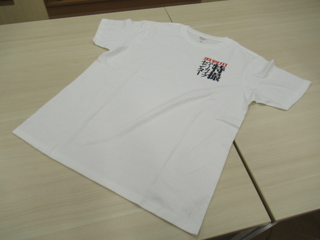 明るい木目調のテーブルの上に広げられた、左胸に「須賀川特撮アーカイブセンター」という黒と赤のロゴがプリントされたシンプルな白い半袖のクルーネックTシャツを斜めの角度から捉えた写真