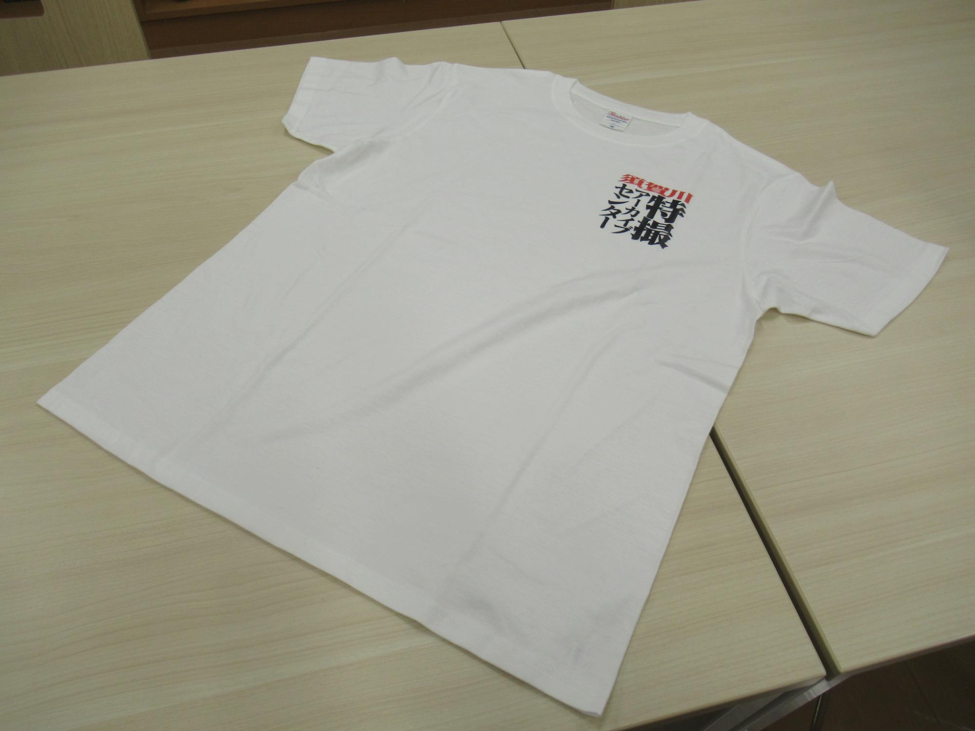 テーブルの上に広げられた、左胸に「須賀川特撮アーカイブセンター」という黒と赤のロゴがプリントされたシンプルな白い半袖のクルーネックTシャツを斜めの角度から捉えた写真