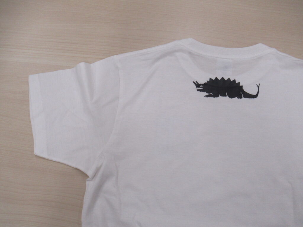 明るい木目調のテーブルに置かれた白い半袖Tシャツの背面をアップで捉えており、首元のリブのすぐ下に、背中に鋭い突起を持ち、頭部から角のようなものが生えた巨大な怪獣のシルエットイラストがワンポイントでプリントされている写真