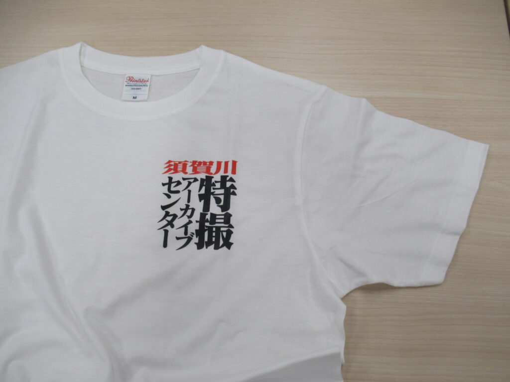 明るい木目調のテーブルに置かれた白い半袖Tシャツの左胸部分をアップで捉えており、赤色で「須賀川」、その下に黒色で「特撮アーカイブセンター」と力強い書体でデザインされたロゴが鮮明に写っている写真