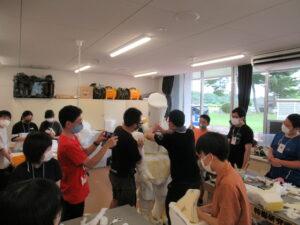 塾生たちが協力しながら白い素材で立体的な造形物を組み立てる作業に取り組み、一人がその様子をスマートフォンで撮影している写真