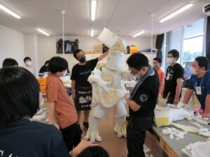 室内で、塾生たちが協力しながら白いスポンジ状のパーツを丁寧に繋ぎ合わせた大きな怪獣の着ぐるみを完成に向けて調整をしている様子の写真