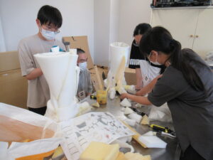 明るい室内で、塾生たちが白いスポンジ状のパーツを丁寧に繋ぎ合わせ、大きな怪獣の足を作っている写真