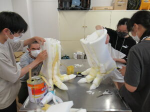 4人の塾生が、屋内で白いスポンジのような素材を繋ぎ合わせ、恐竜の足のような大きな造形物を2つ制作している写真