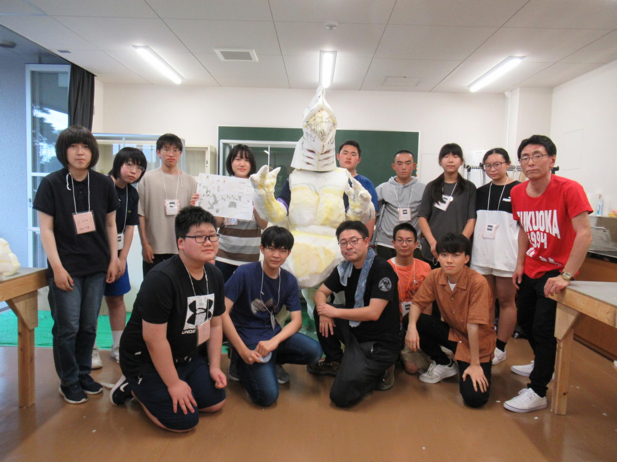 屋内で白い怪獣の着ぐるみを囲み、複数の塾生たちと講師が集まって記念撮影をしている写真