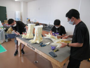 塾生2名と男性が作業台の上で、カッターなどの工具を使いながら、ウレタン素材と思われる怪獣の巨大な足や爪のパーツを製作している写真