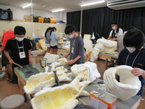 室内で、数名の塾生たちが、作業台の上に広げられたウレタン素材の白い怪獣のパーツをカッターで削ったり接着したりして、巨大な着ぐるみの各部位を作り上げている写真