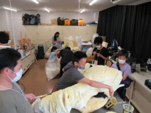 明るい室内で数人の若者が協力し合い、大きな黄色い模型のような物体に布を被せたり接着剤を塗ったりして制作活動に励んでおり、背景の棚には機材や荷物が並んでいる、作業風景を捉えた写真