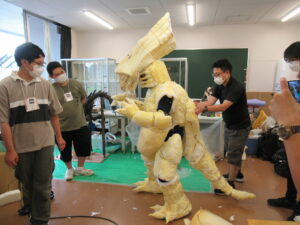 黄色い発泡スチロールのような素材で作られた巨大な怪獣が中央に立ち、講師が調整をし、その周りで塾生が遠目で確認している様子の写真