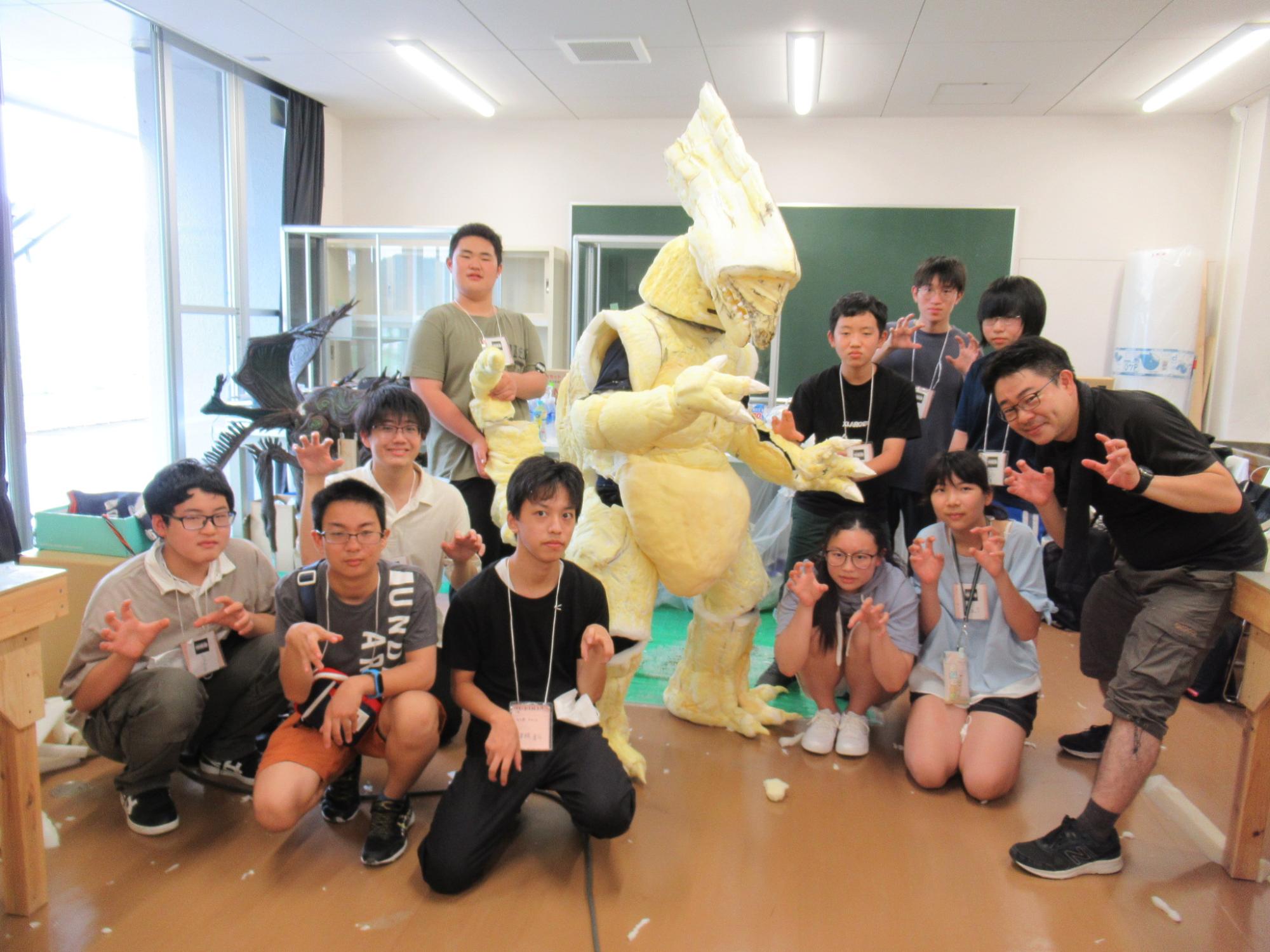 教室の黒板の前に、手作りの巨大なスポンジを付けた怪獣の着ぐるみを囲んで集まった複数の男女の塾生たちが、両手を爪のように立てる怪獣のポーズをしながらカメラを見つめている集合写真