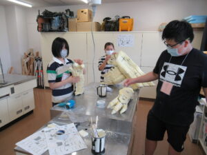 室内で若者たちが、淡い黄色の素材で作られた巨大な怪獣の鉤爪のような造形物や複雑な形状のパーツを机に並べ、手作業で細かな調整や組み立てを行っている様子の写真