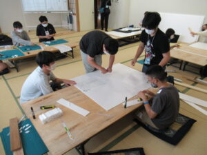 畳敷きの和室で、塾生たちが机を囲み、大きな白いボードやカッター、定規などの道具を用いて真剣な表情で工作や設計の作業に取り組んでいる様子の写真