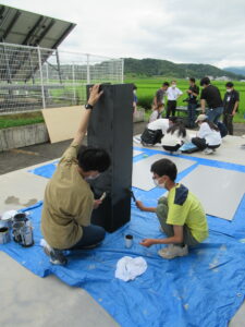 屋外で、青いビニールシートの上に置かれた背の高い黒色の立体的な構造物に、二人の塾生がしゃがみ込みながら刷毛を使って丁寧に黒い塗料を塗り重ねている写真