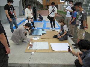 屋外のコンクリート床の上で、数人の塾生たちがしゃがみ込み、段ボールの上に白いシートを広げて大型の制作物を組み立てる作業を周囲で見守る仲間たちと共に進めている様子の写真