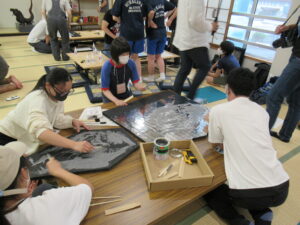 畳敷きの和室で、数名の塾生たちが長机を囲み、黒いボードの上に細かな作業をして進めている様子の写真