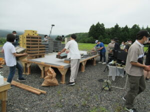 屋外の砂利敷きの広場で、木製の台座の上にビルや民家の精巧なミニチュア模型を慎重に配置して街並みを作り上げている写真